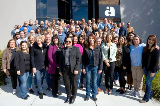 A large, diverse group of smiling professionals poses together on concrete steps outside a modern office building. A black sign with a stylized white letter "a" logo is visible on the wall behind them.
