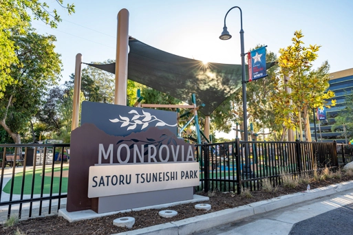 A park entrance sign reads "Monrovia Satoru Tsuneishi Park" against a backdrop of a playground with a black shade sail under bright sunlight.