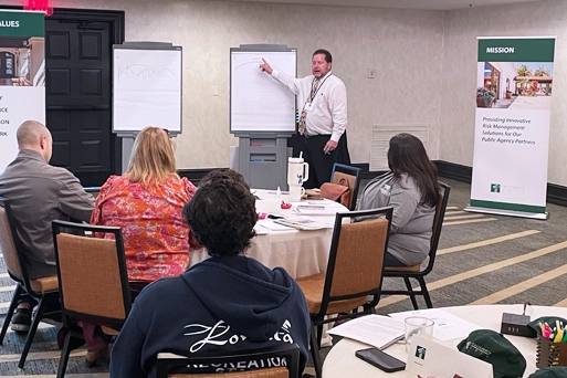 A man in a white button-down shirt and tie points to a whiteboard while leading a presentation for an audience seated around a table in a conference room.