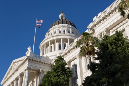 A low-angle, sunlit view shows the white dome and facade of the California State Capitol building in Sacramento against a clear, deep blue sky.